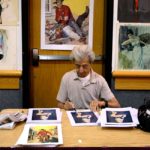 Ernest Chiriacka, one of Pulpcon’s guests of honor, signs prints of one of his pin-up paintings, next to a stack of pulp cover prints, at his table in the dealers’ room.